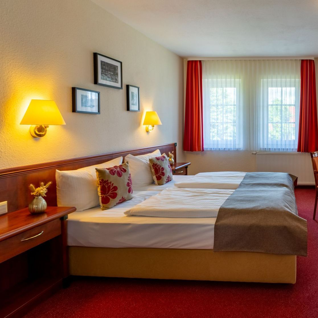 Hotelzimmer mit zwei Einzelbetten, rotem Teppich und zwei Fenstern mit roten Vorhängen.