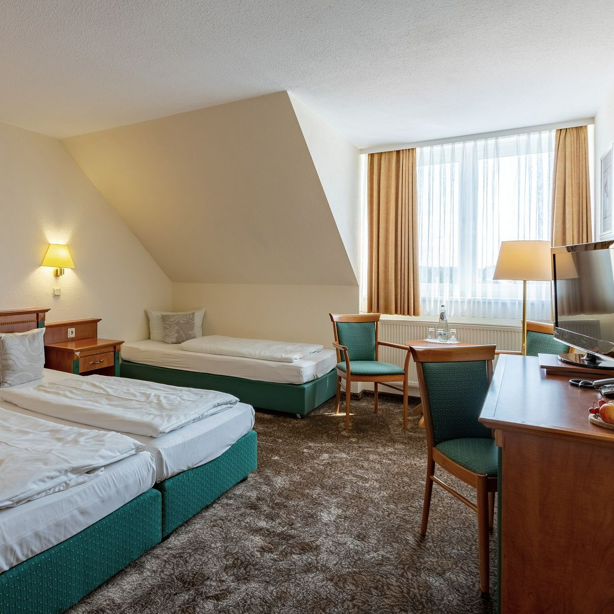 Hotelzimmer mit zwei Einzelbetten, einem Einzelbett, Schreibtisch und Stühlen. Beige Wände, brauner Teppich.