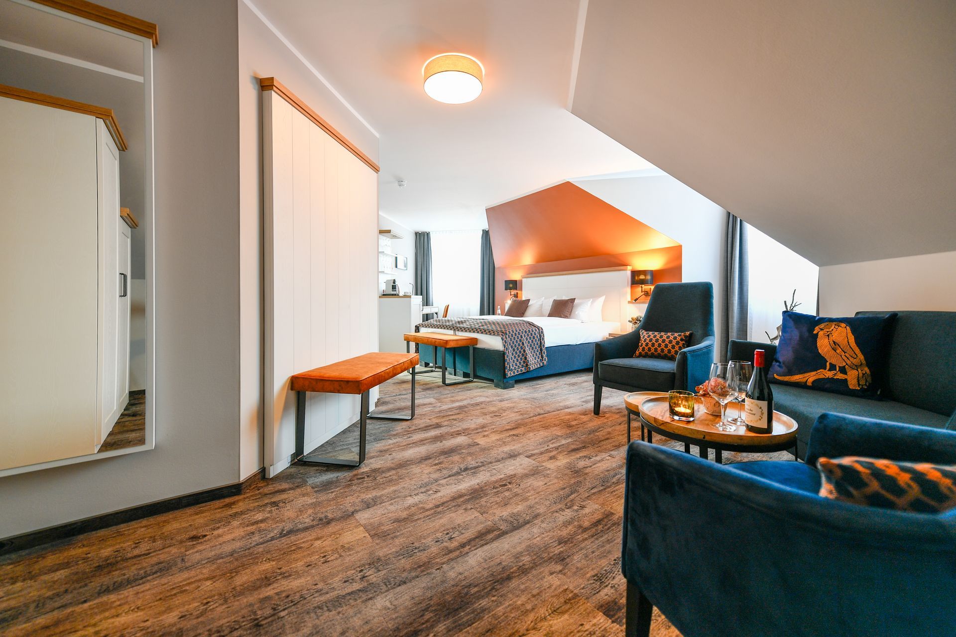 Hotelzimmer mit blauen Samtmöbeln, orangefarbenen Akzenten und einem Bett unter einer Dachschräge.