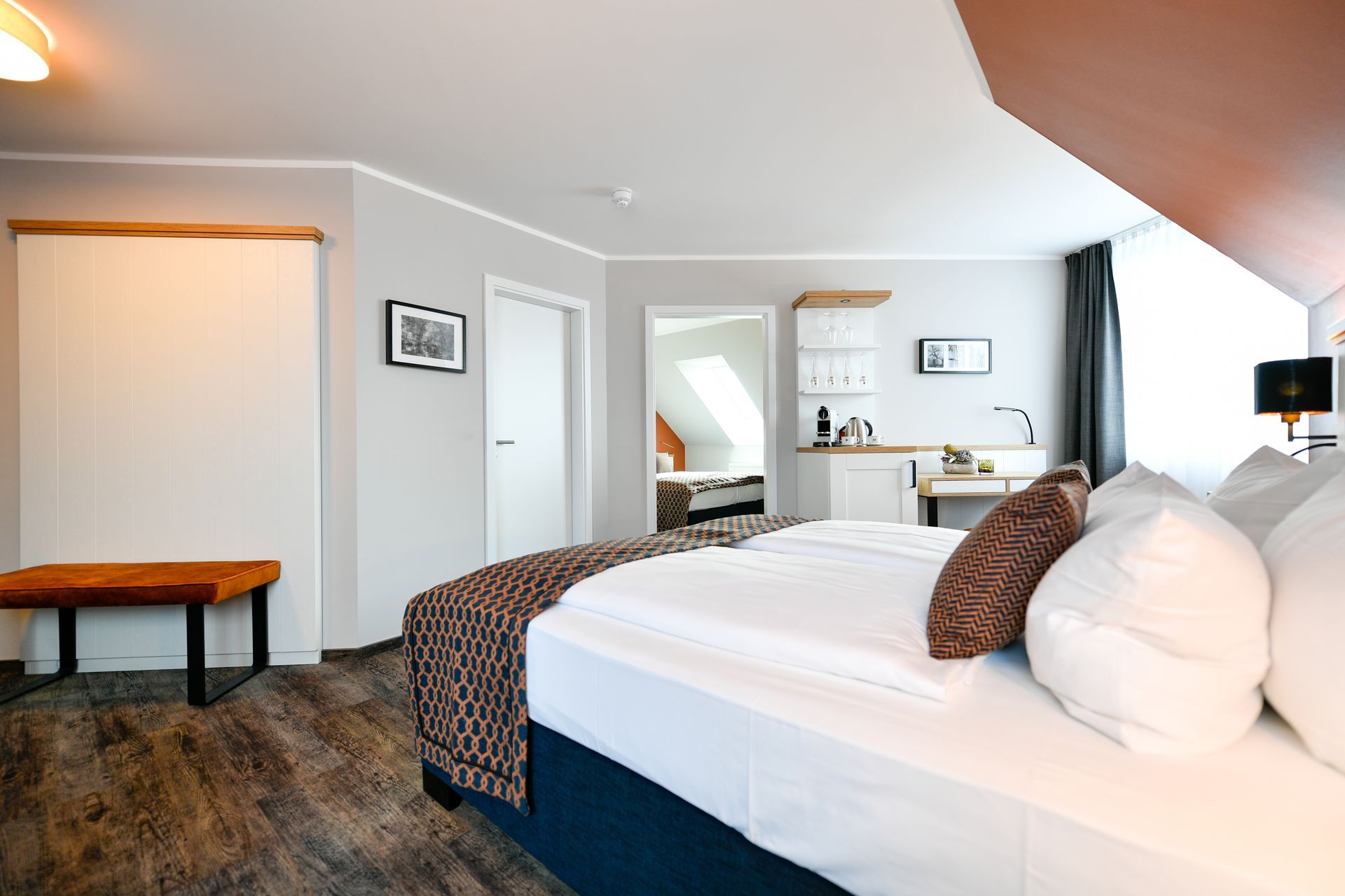 Schlafzimmer mit Bett, Bank, Arbeitsbereich und kleiner Küchenzeile. Graue Wände, Holzboden und weiße Bettwäsche.