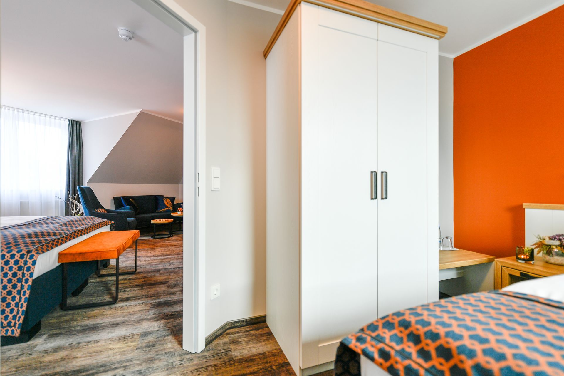 Weißer Kleiderschrank und offene Tür zum Wohnbereich, orange Akzentwand, zwei Betten mit gemusterten Decken.