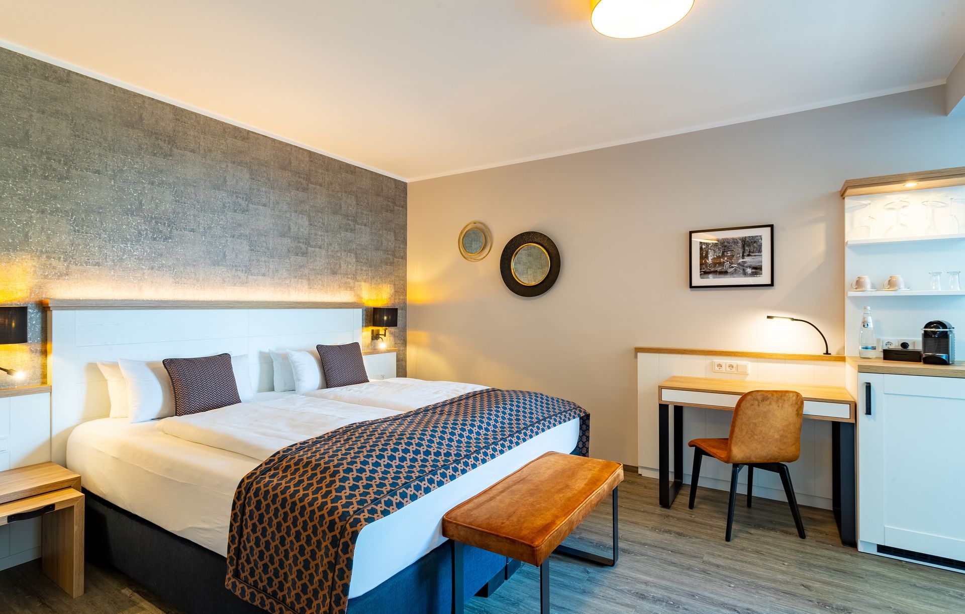 Hotelschlafzimmer mit Kingsize-Bett, Schreibtisch, Bank und grau strukturierter Wand.