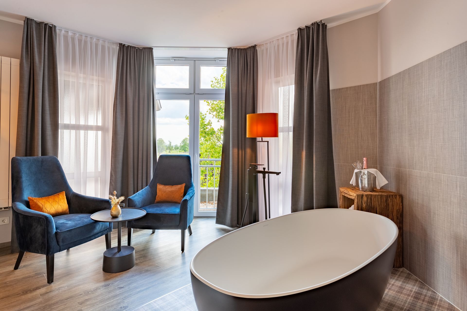 Hotelzimmer mit blauen Sesseln, einer Badewanne und einem Fenster mit Balkon und Blick ins Grüne.