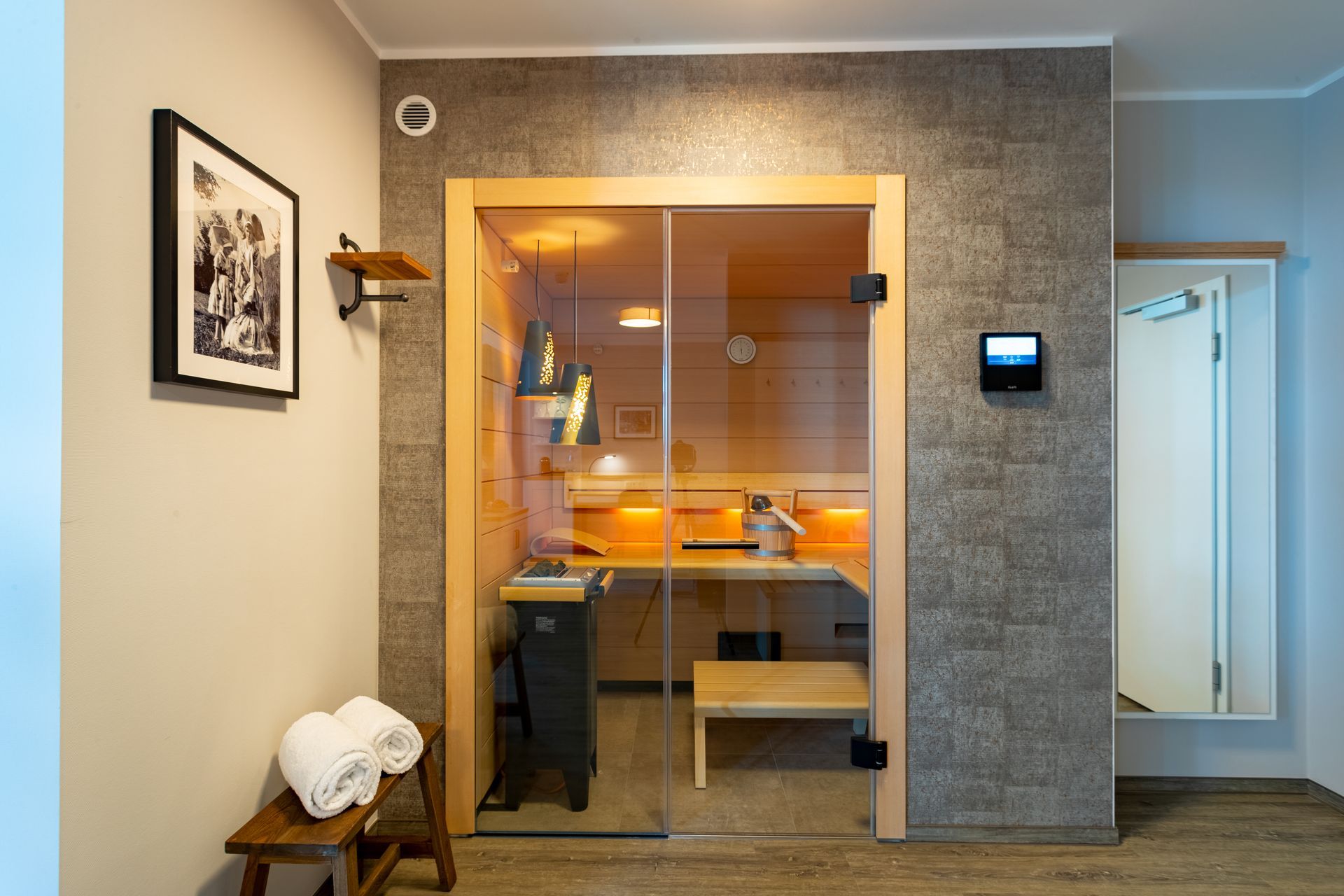 Sauna mit Glastür, Holzbank und Handtüchern auf einem Hocker. Graue Steinwand.