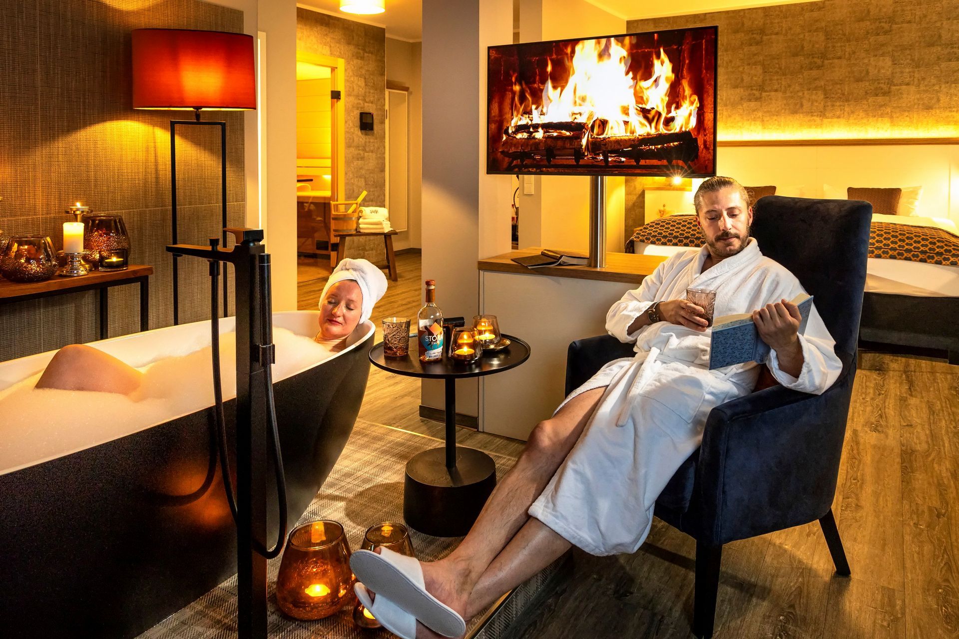 Ein Paar entspannt in einer Luxussuite: Frau in der Badewanne, Mann im Bademantel beim Lesen, Kaminfeuer im Fernsehen.