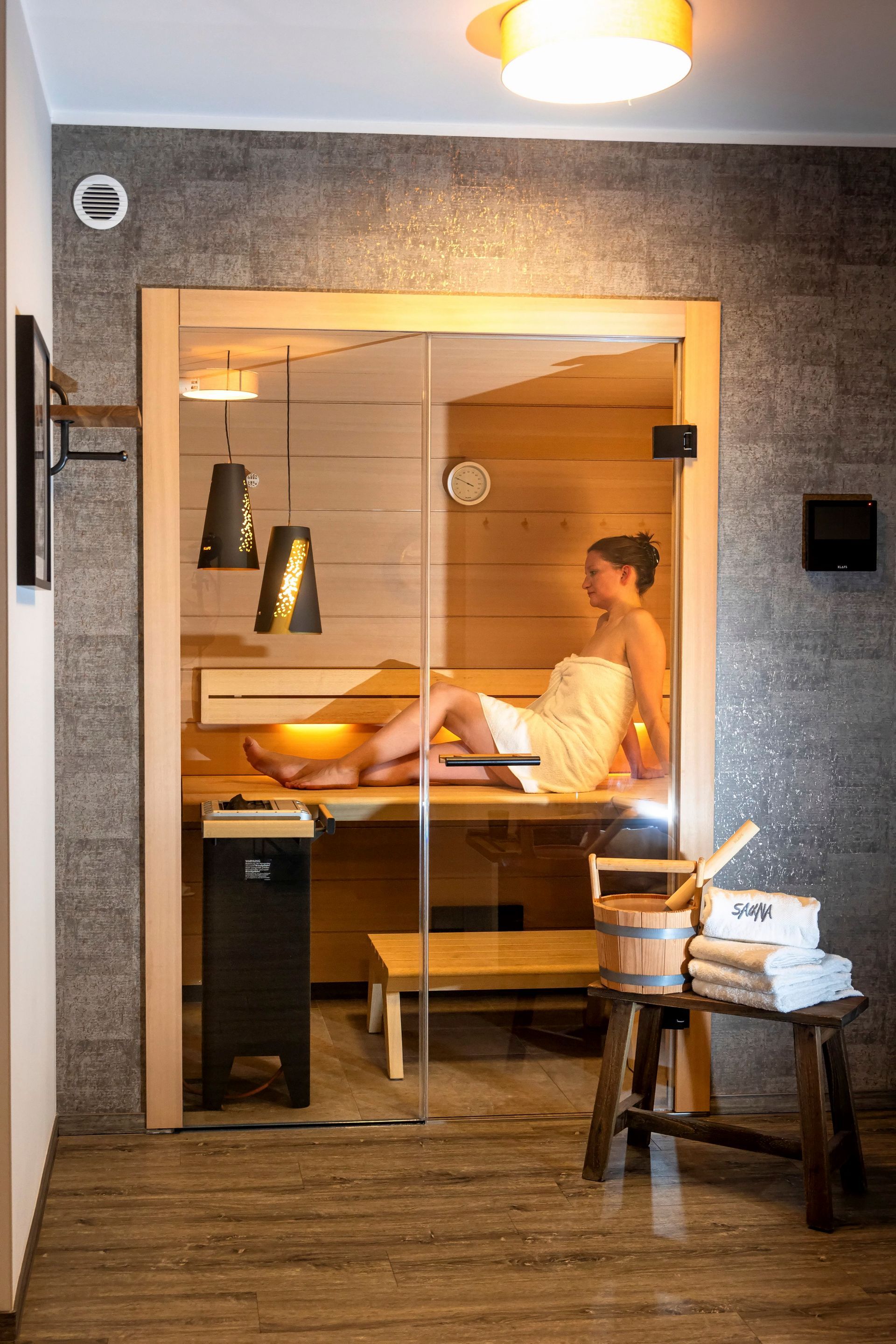 Frau entspannt in einer holzgetäfelten Sauna mit Glastür, Leuchte, Handtuch und Holzhocker.