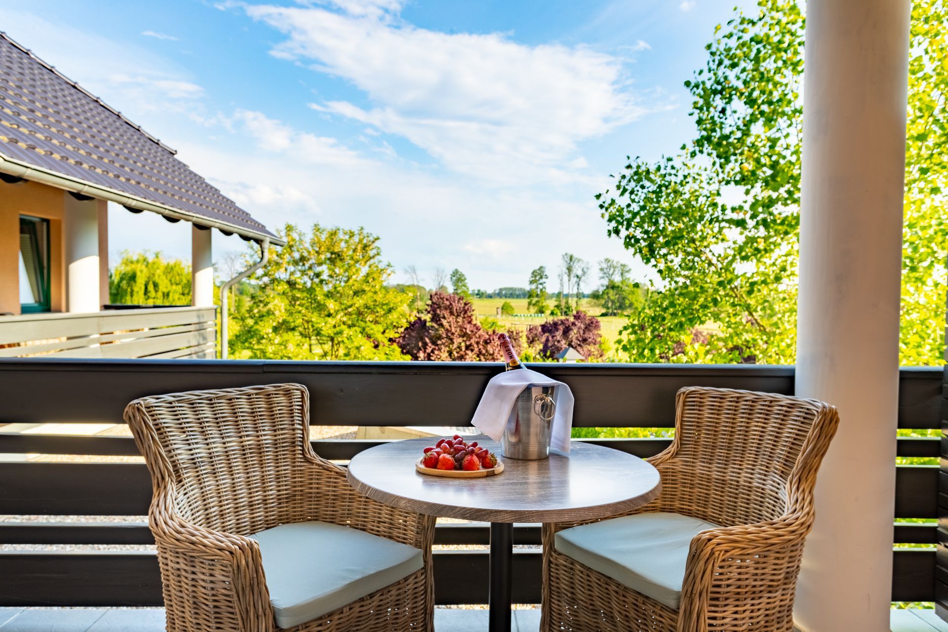 Balkon mit Korbstühlen, Tisch mit Erdbeeren und Wein; Blick auf grüne Landschaft, blauer Himmel.