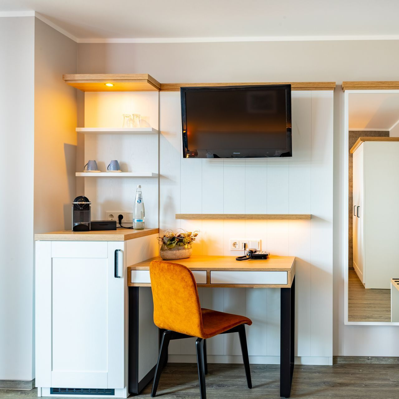 Ein geräumiges Zimmer mit einem Schreibtisch, einem orangefarbenen Stuhl, einem Fernseher und weißen Schränken.