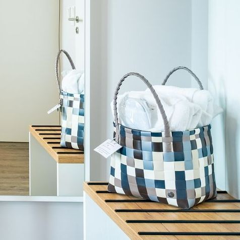 Geflochtener Korb in Blau, Grau und Weiß, gefüllt mit weißem Stoff, auf einem Holzregal mit Spiegel.