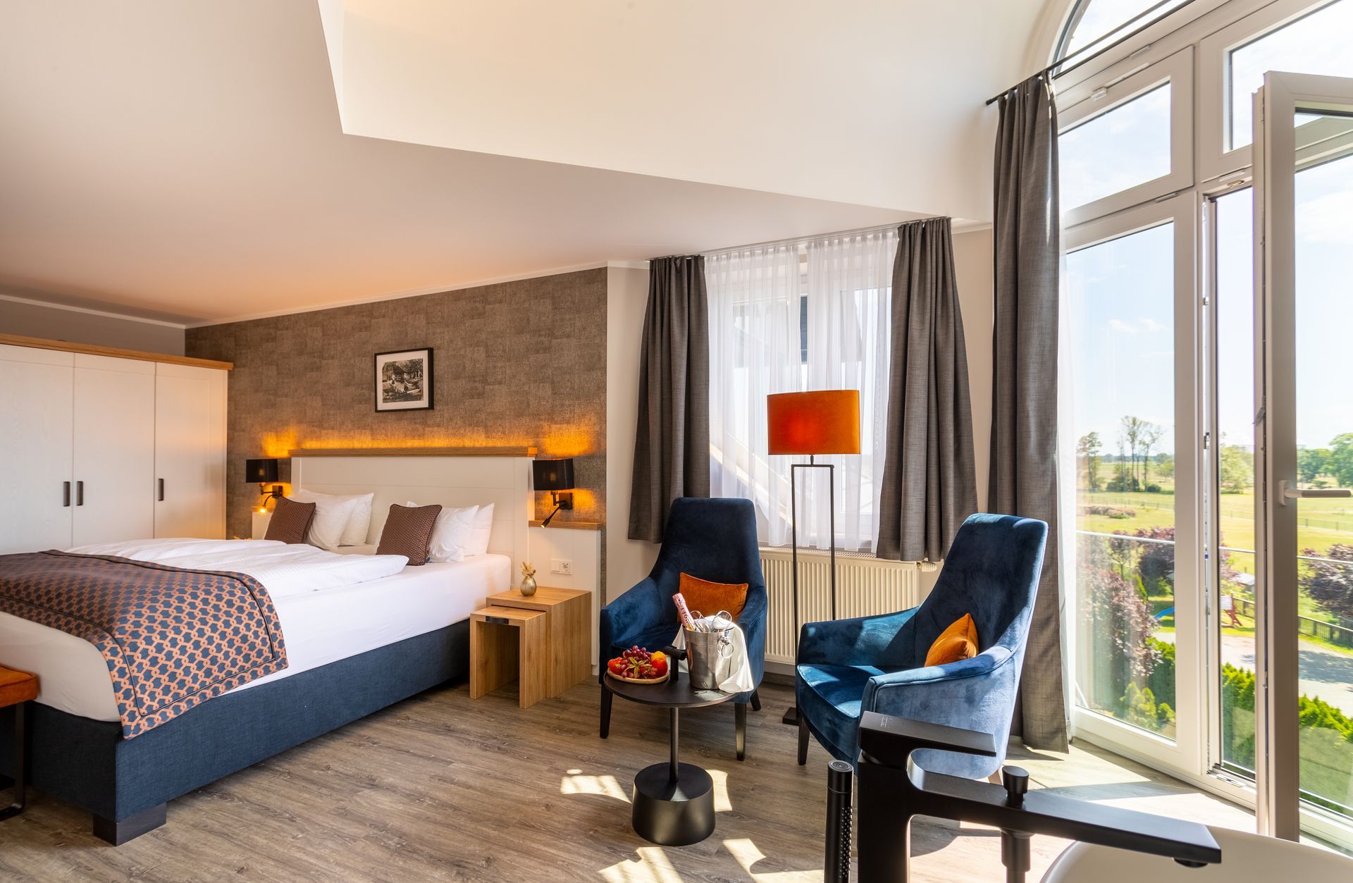 Hotelzimmer mit Bett, zwei blauen Sesseln, großem Fenster und orangefarbener Lampe.