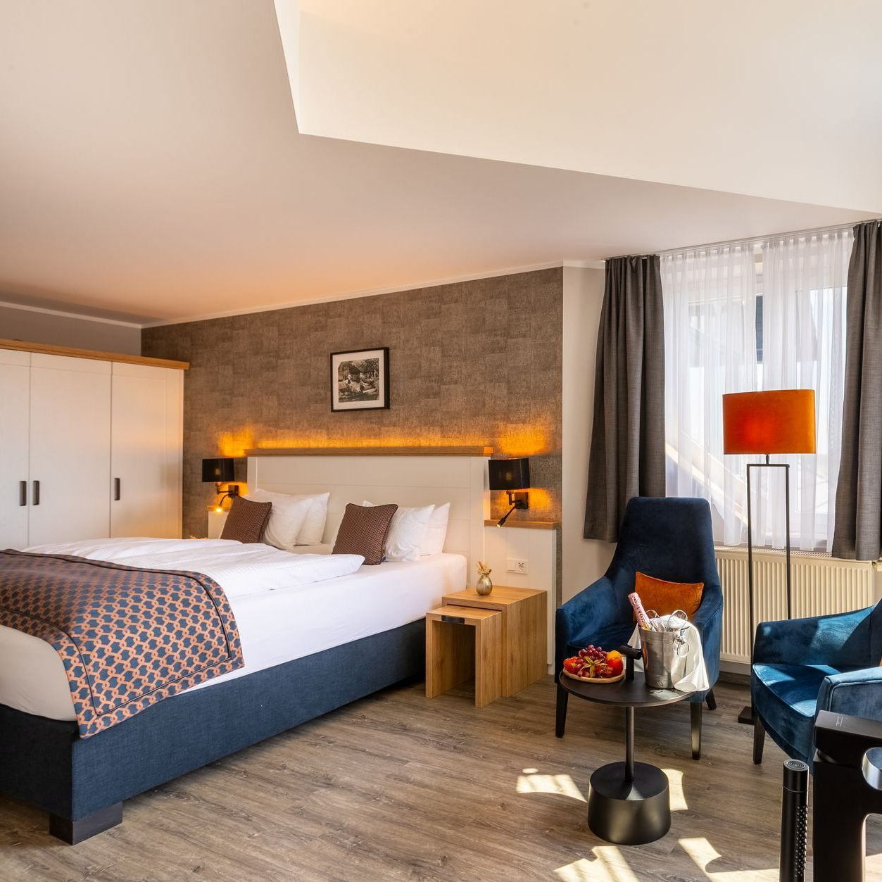 Hotelzimmer mit Bett, Sesseln, Beistelltisch und einem großen Fenster mit Vorhängen.