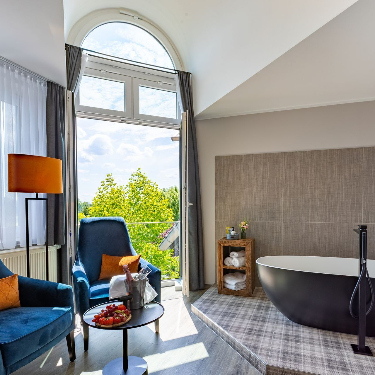 Eine luxuriöse Hotelsuite mit blauen Sesseln, einer schwarzen Badewanne und einem Balkon mit Blick auf Bäume.
