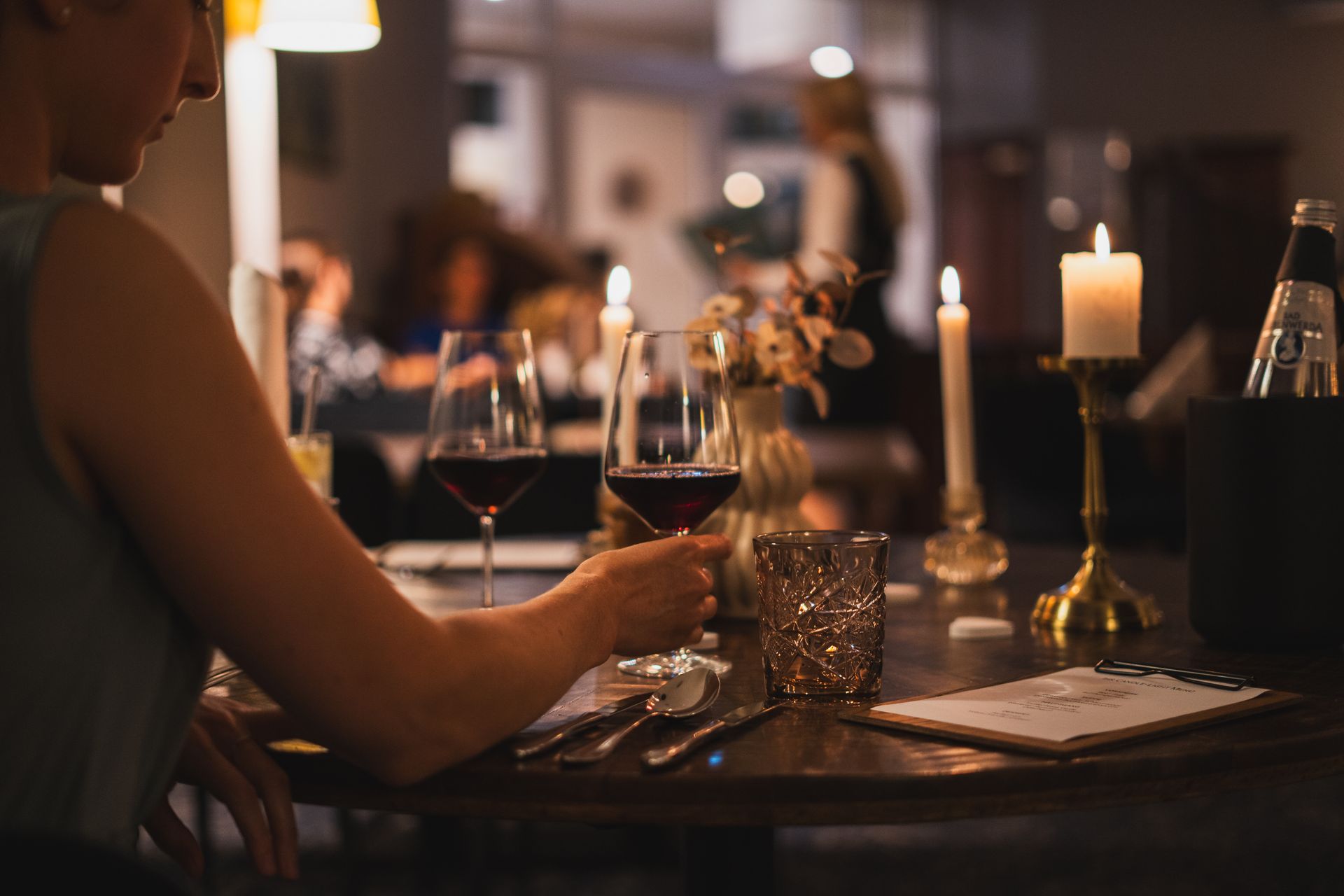 Frau am Restauranttisch mit einem Glas Rotwein in der Hand; brennende Kerzen, andere Gäste im Hintergrund.