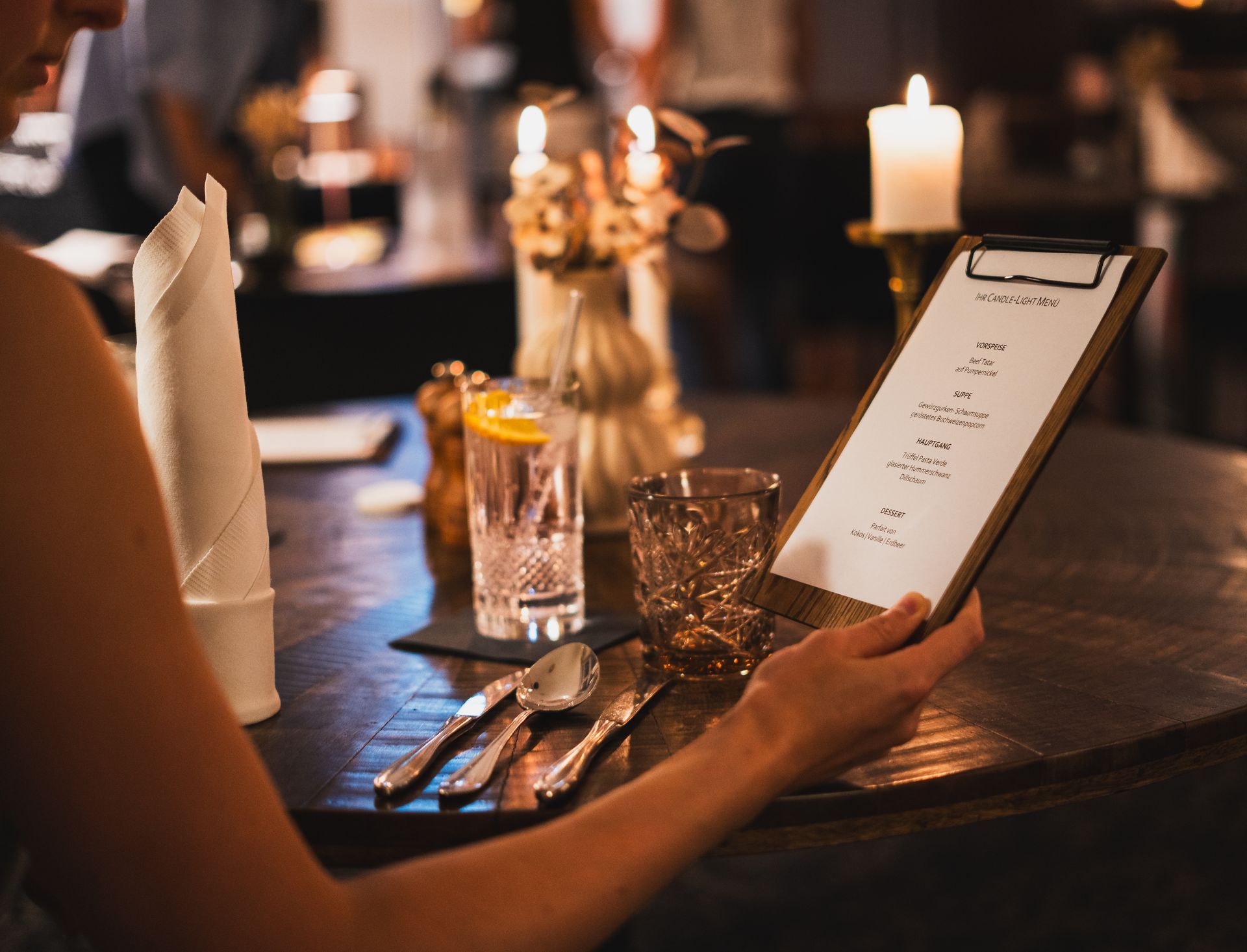 Frau liest an einem Restauranttisch bei Kerzenlicht eine Speisekarte und hält ein Klemmbrett, Getränke und Besteck in der Hand.