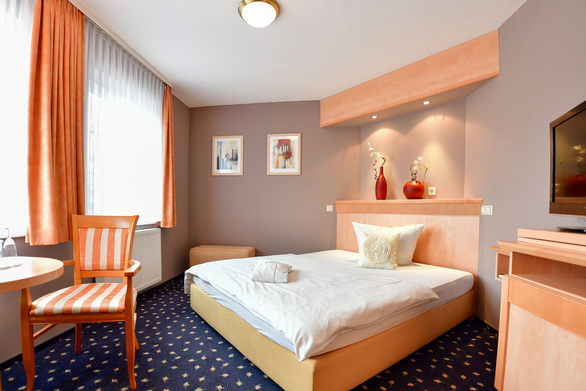 Hotelzimmer mit Bett, Stuhl, Tisch und Fenster mit orangefarbenen Vorhängen; Blumenkunst und Vasendekor.