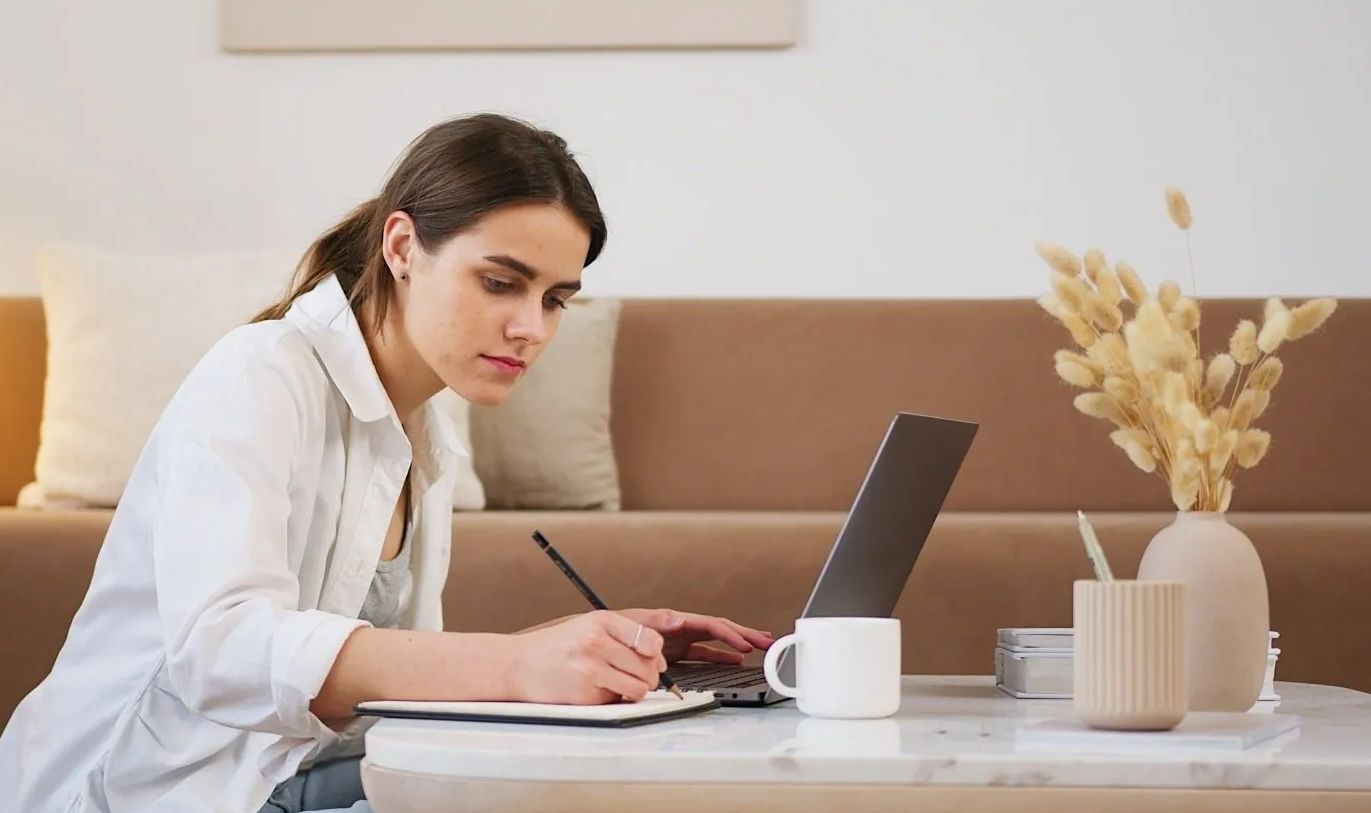 Frau im weißen Hemd arbeitet am Laptop, schreibt in ein Notizbuch, sitzt mit einer Kaffeetasse an einem Couchtisch.