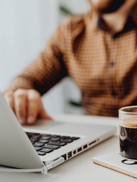 Person mit Laptop, braun kariertem Hemd, Kaffeetasse auf dem Schreibtisch.