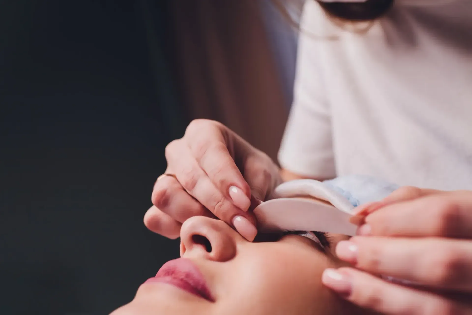 Mujer recibiendo extensiones de pestañas; técnico colocando almohadillas para los ojos.