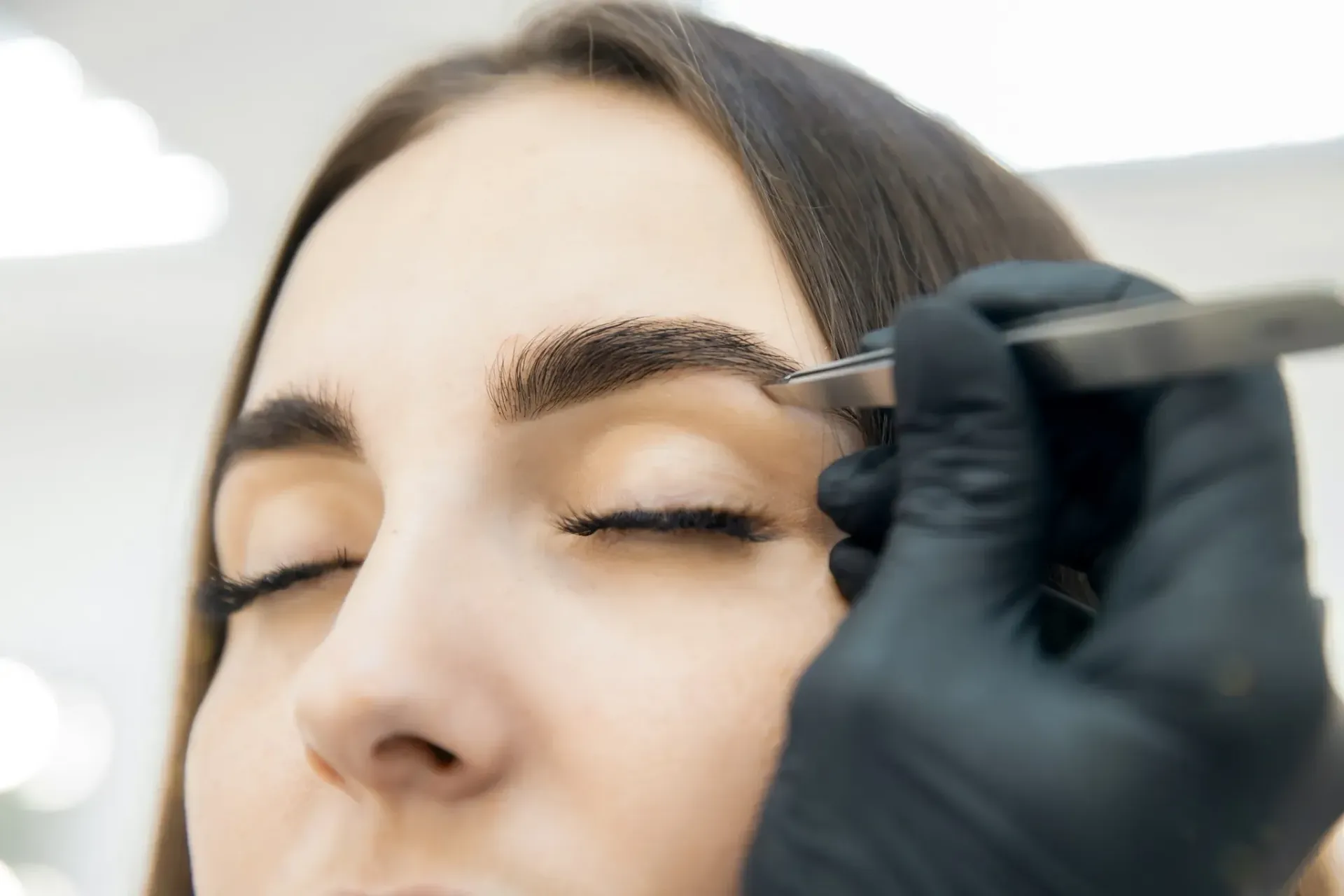 Persona depilándose las cejas con pinzas, primer plano. Ojos cerrados, mano enguantada.