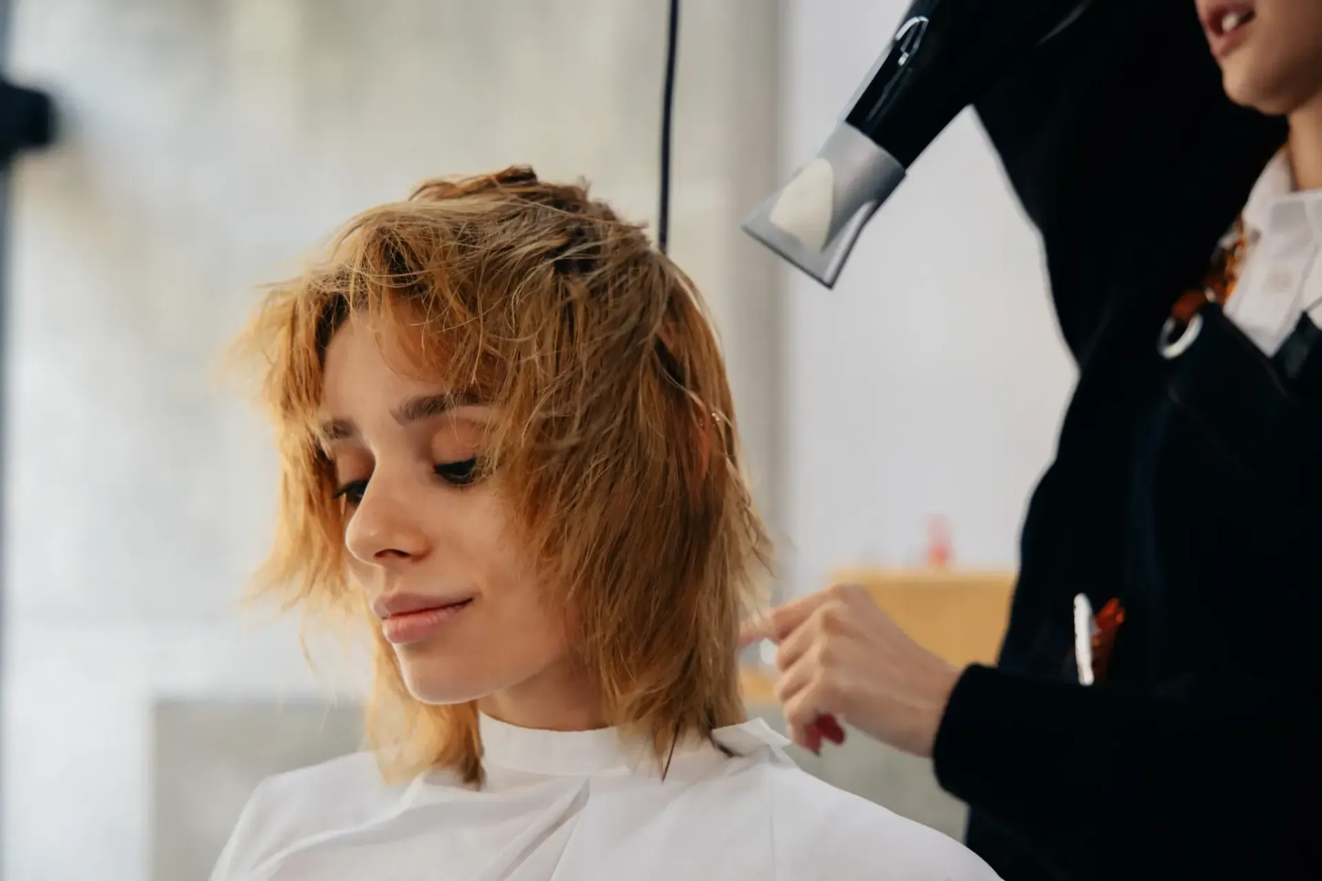 Mujer secándose el cabello en el salón; estilista sosteniendo un secador de pelo.