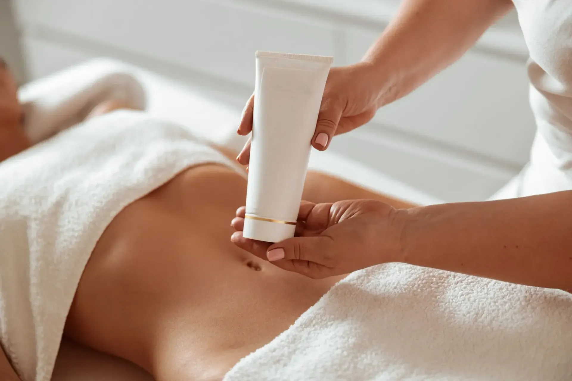 Persona aplicando crema de un tubo en el abdomen de otra persona, ambas en una mesa de masaje.