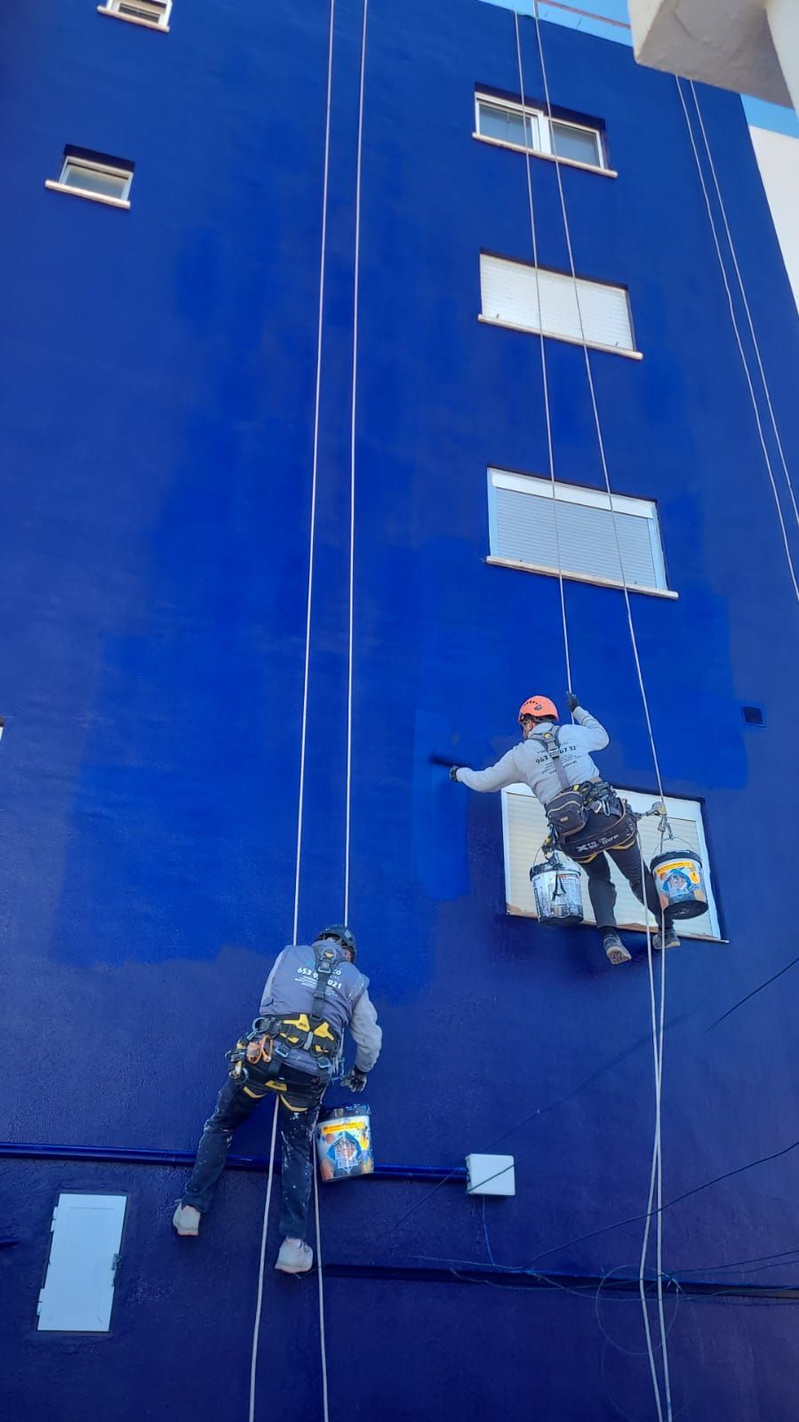 Dos hombres están pintando el costado de un edificio azul.