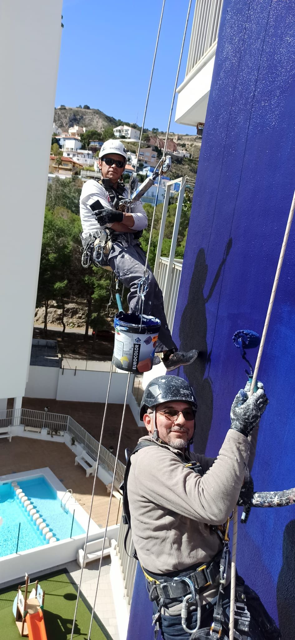 Dos hombres están pintando una pared azul de un edificio.
