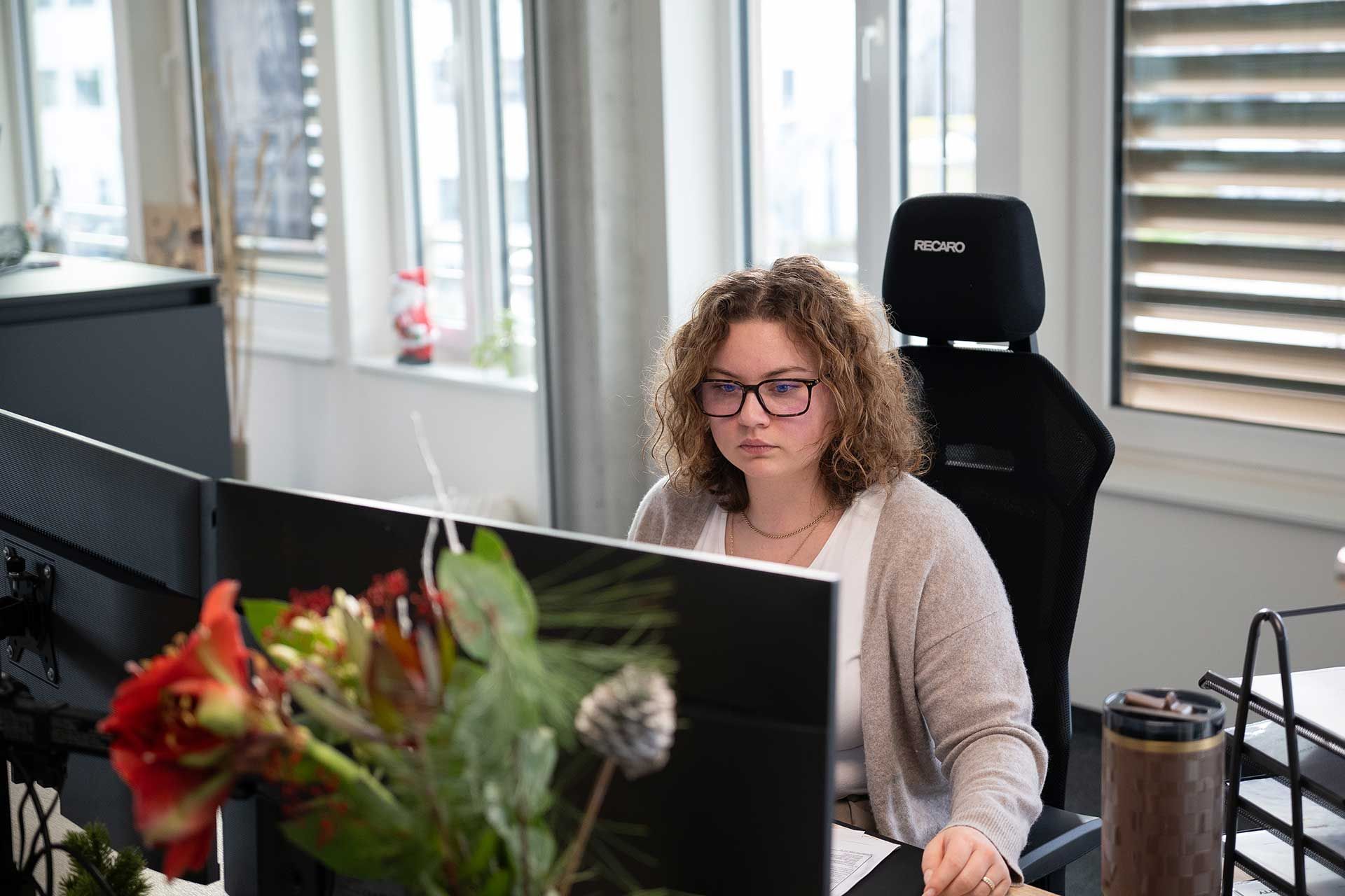 Frau mit Brille arbeitet an einem Computer in einem Büro; im Vordergrund sind Pflanzen zu sehen.
