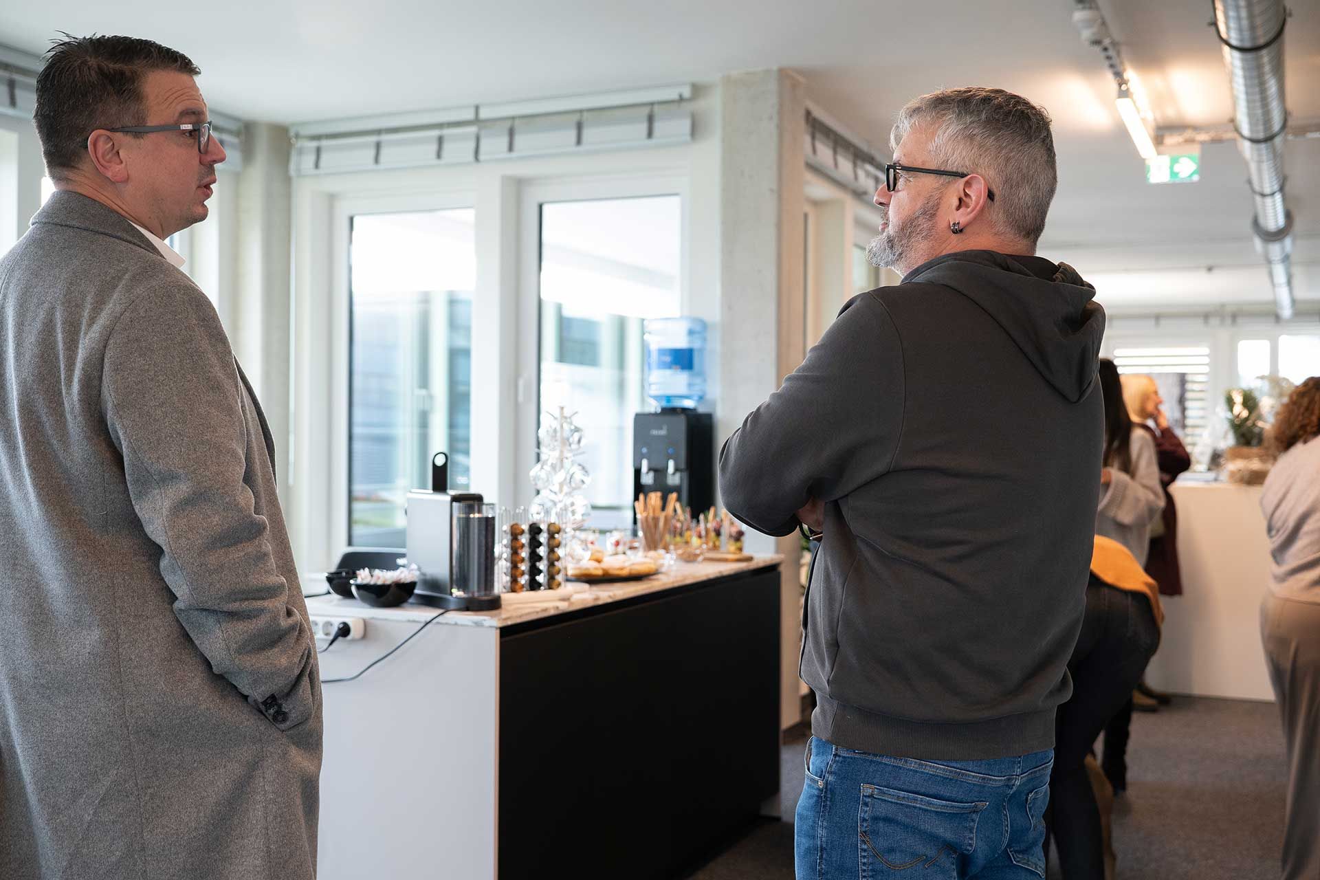Zwei Männer unterhalten sich in der Nähe einer Kaffeestation in einem hellen Büro.
