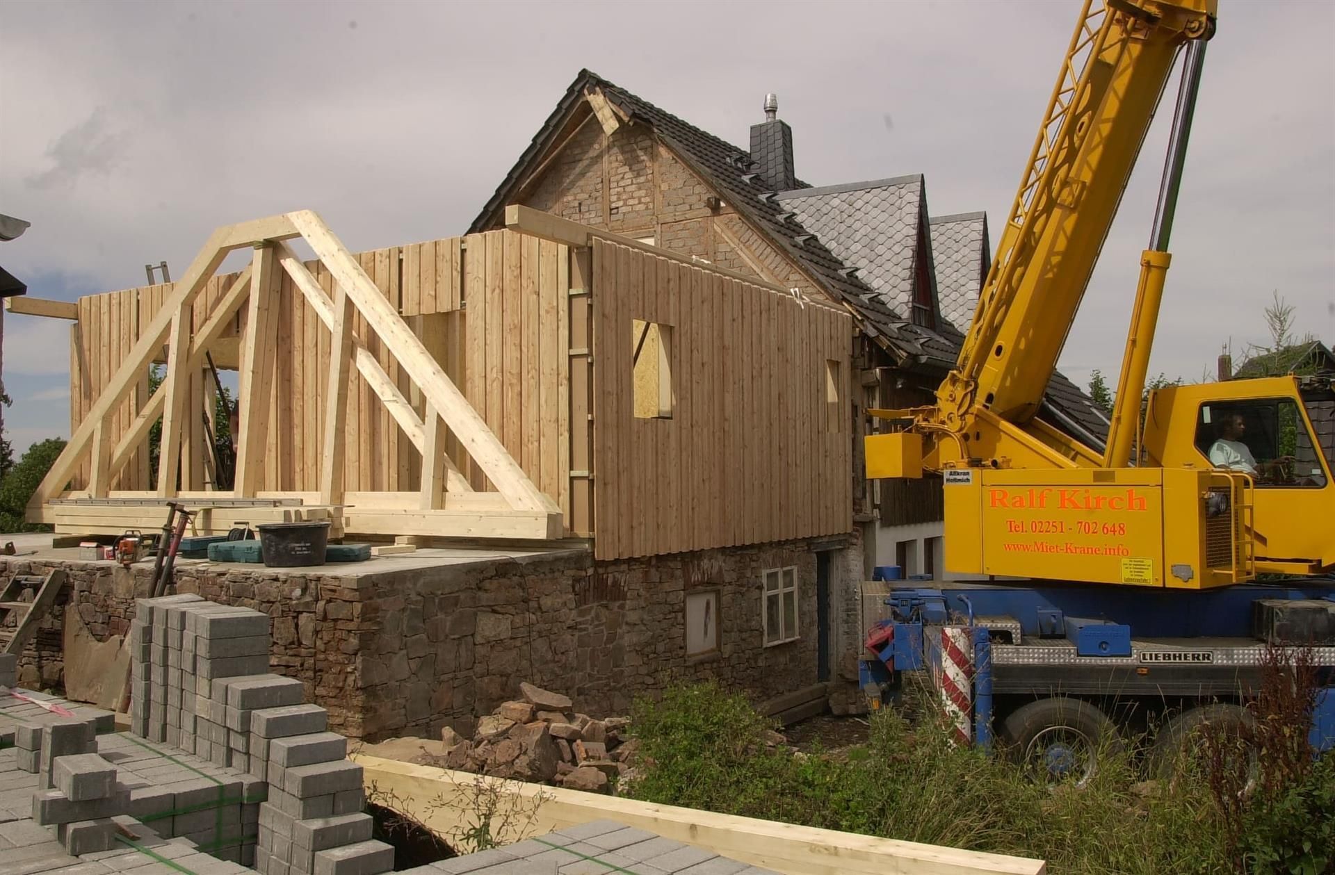 Ein gelber Kran hebt eine Holzkonstruktion vor einem Haus an