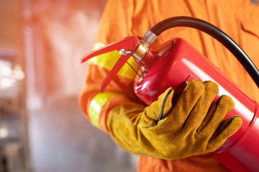 Main gantée de jaune tenant un embout d'extincteur rouge dans une scène enfumée.