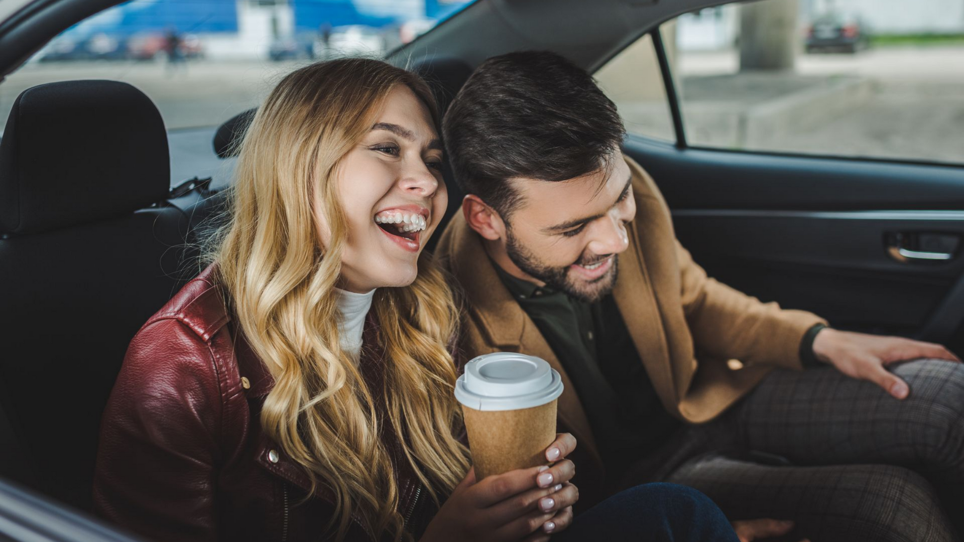 Une femme rit, un café à la main, à côté d'un homme souriant dans une voiture.