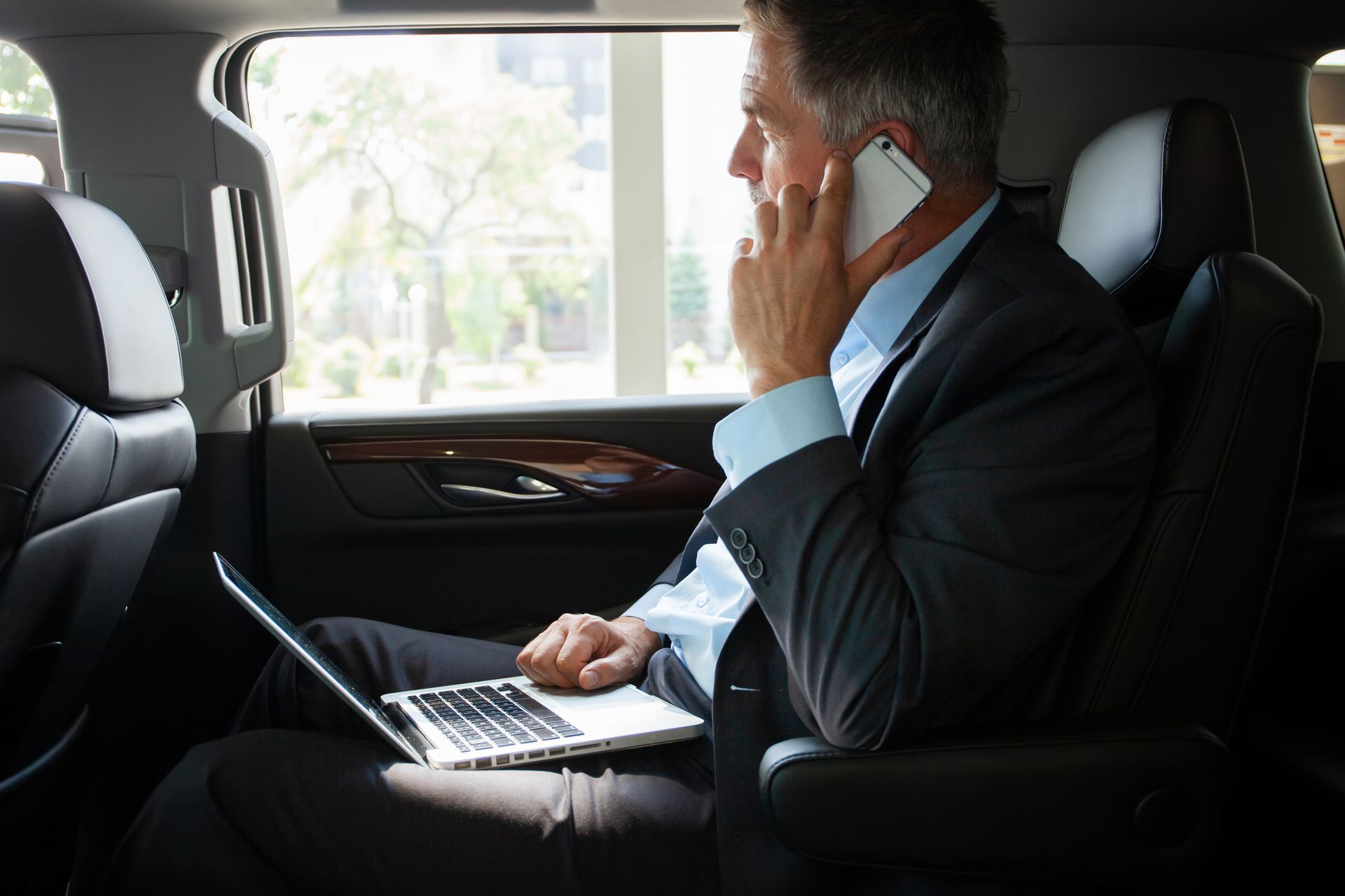 Un homme en costume, au téléphone, utilise un ordinateur portable sur la banquette arrière d'une voiture.