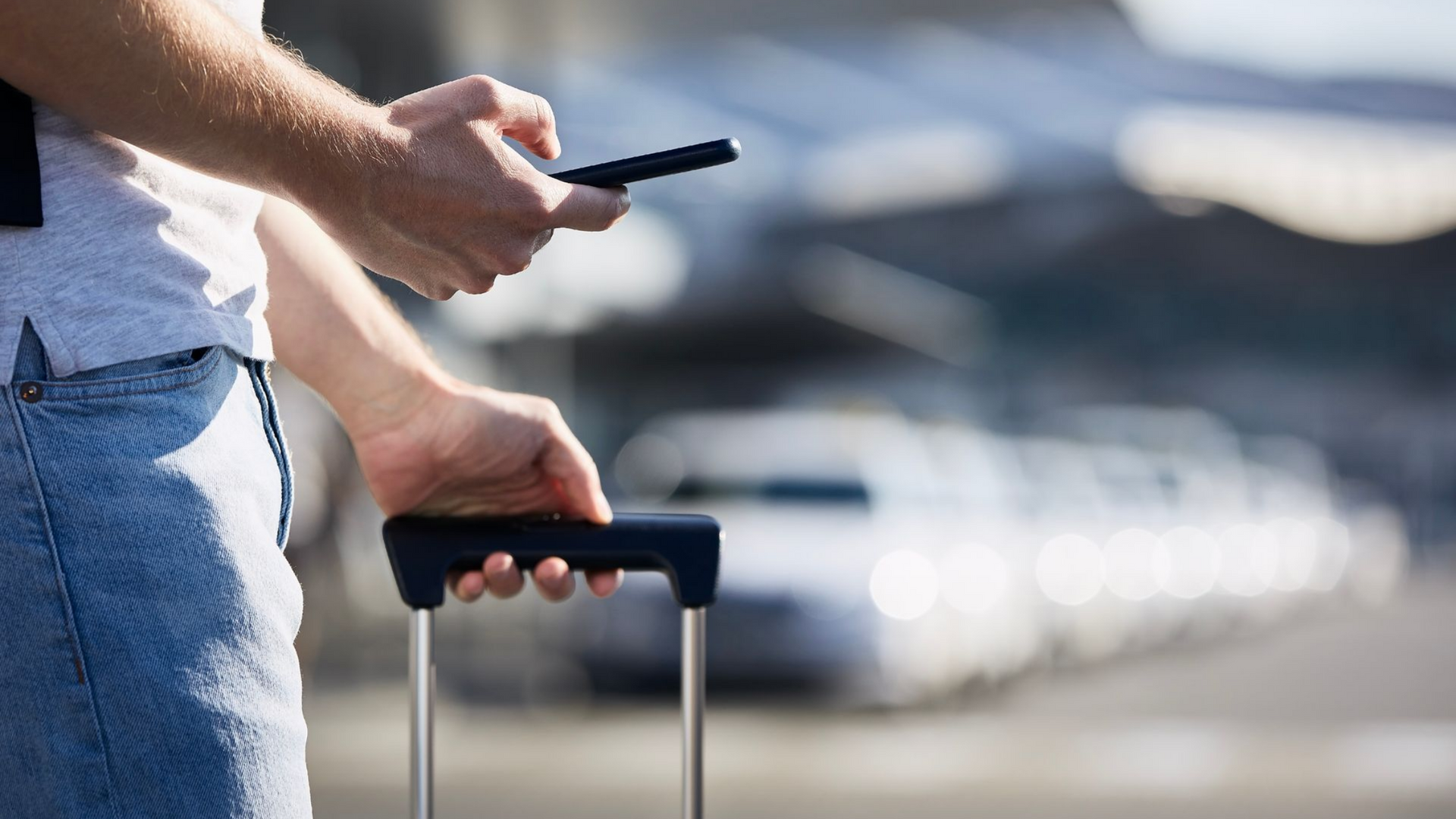 Une personne utilise un smartphone, tenant la poignée de sa valise devant un aéroport.