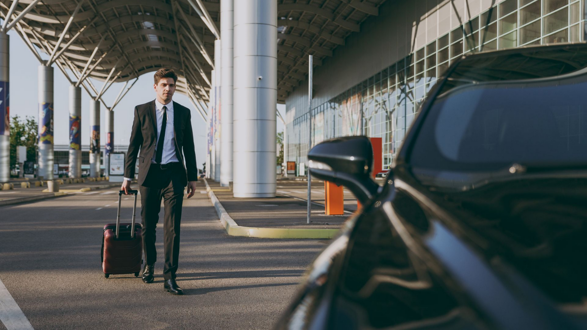 Un homme en costume avec des bagages se dirige vers une voiture noire à l'extérieur de l'aéroport.
