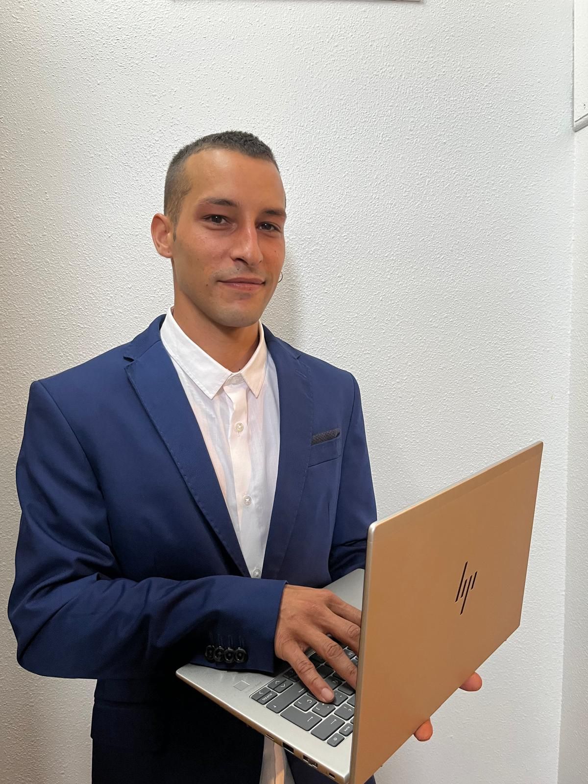 Hombre con traje azul, sosteniendo una computadora portátil, sonriendo, de pie junto a una pared blanca texturizada.