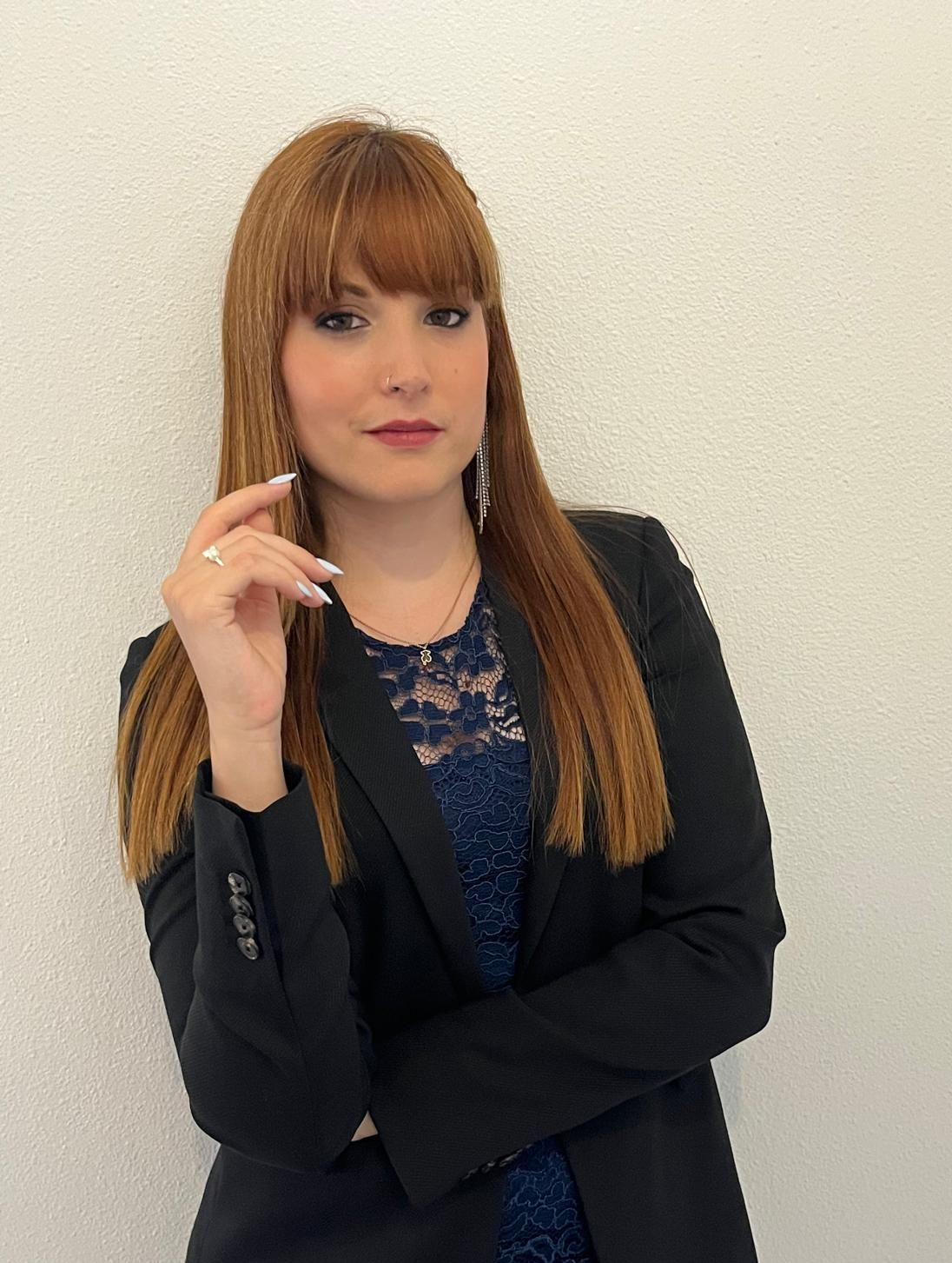Mujer de cabello rojo, vestida con un blazer negro sobre un top de encaje azul, apoyada contra una pared blanca.