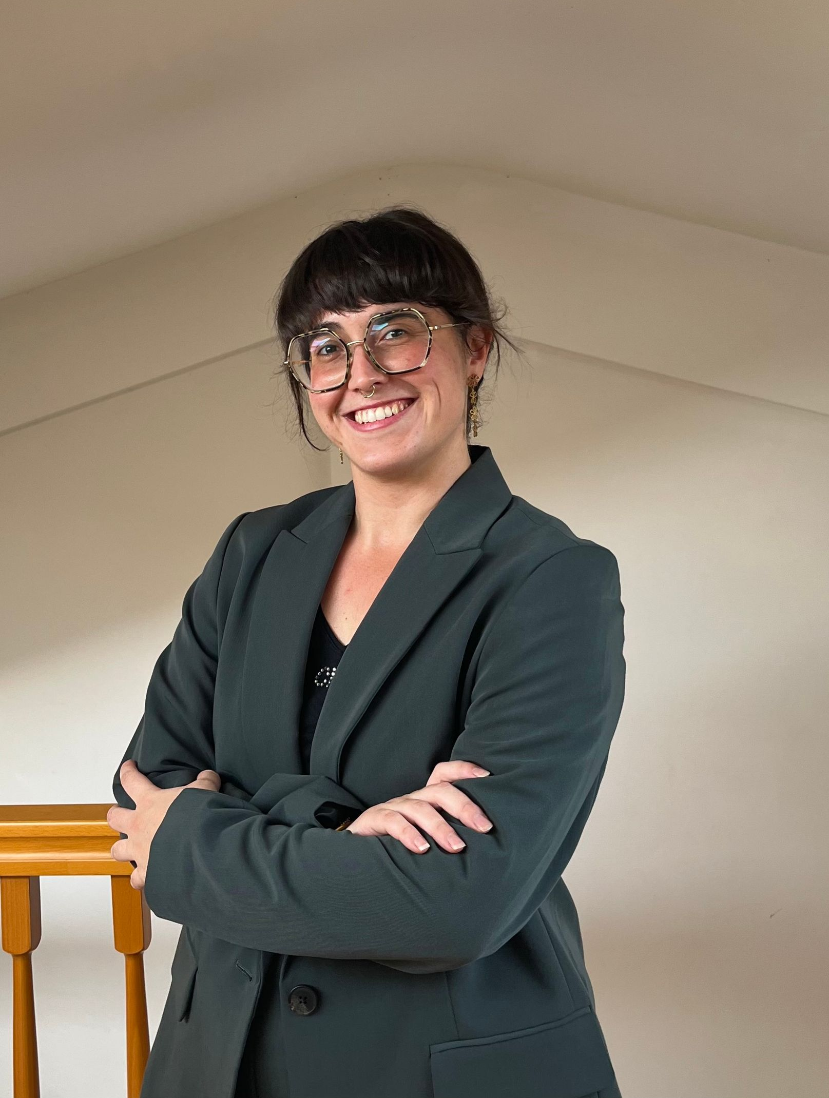 Mujer con un blazer verde oscuro y gafas, con los brazos cruzados, sonriendo en una habitación con escalera.