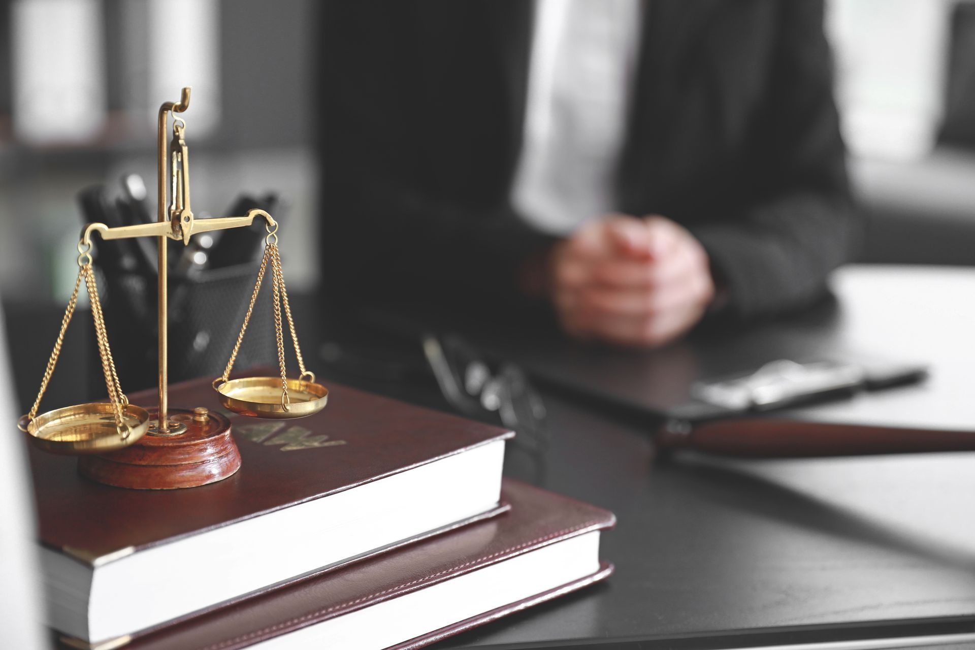 Balanza de la justicia sobre libros delante de una persona con traje, probablemente un abogado, fondo borroso.