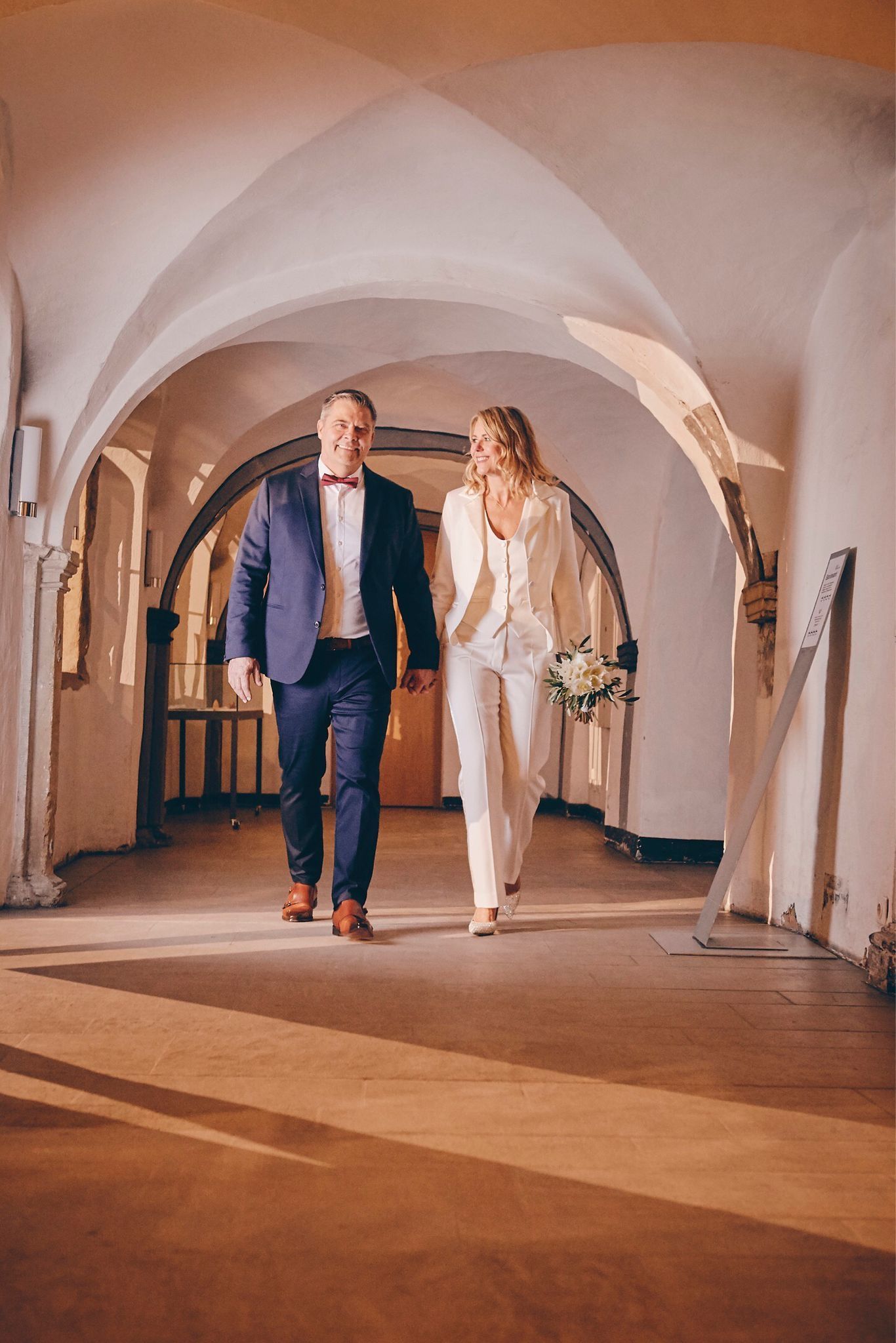 Ehepaar läuft der Kamera entgegen, Braut in weißem Anzug, Bräutigam in blauem Anzug