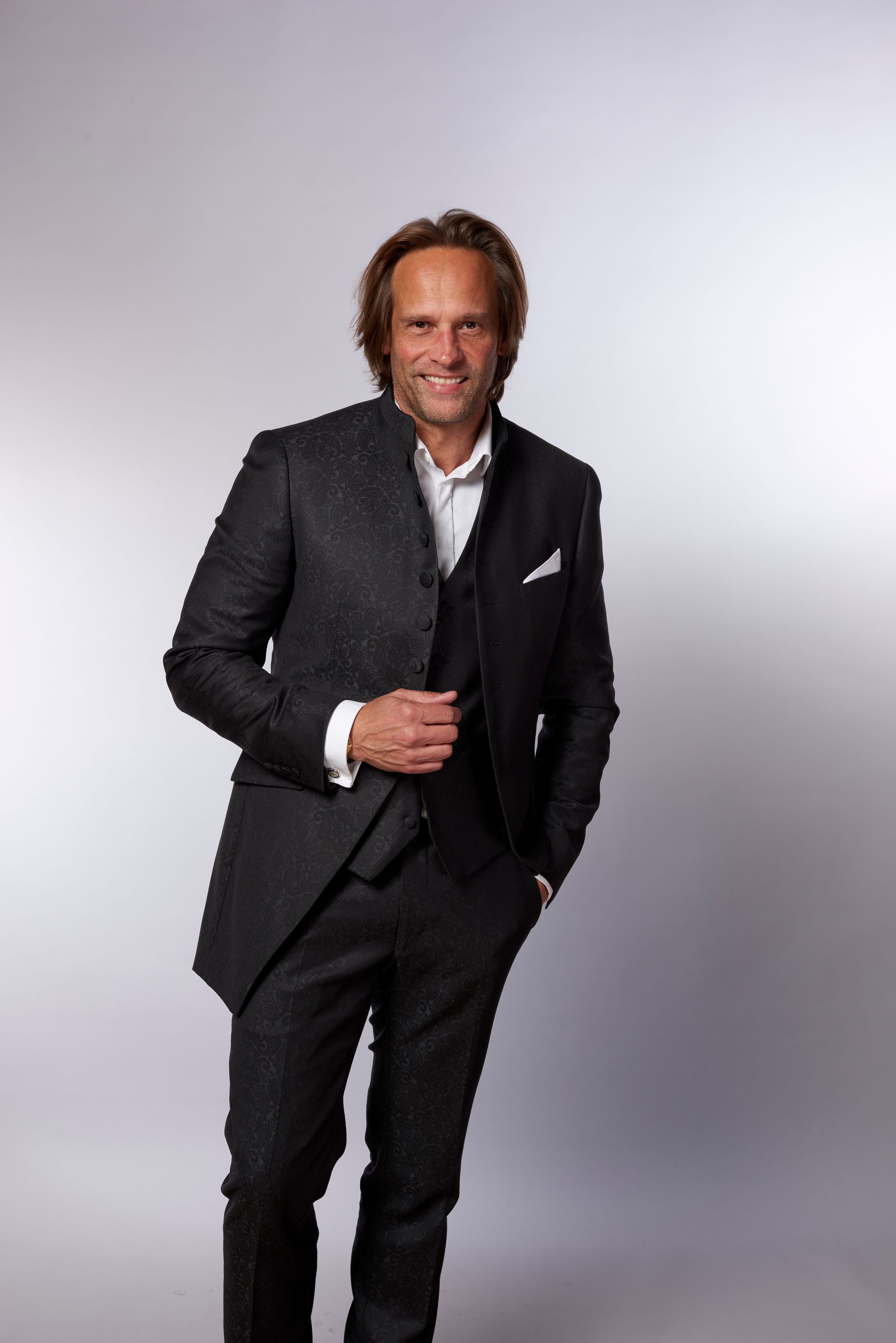 Mann im dunklen Anzug, weißes Hemd, lächelnd, Hand in der Tasche, vor einem Farbverlaufshintergrund.