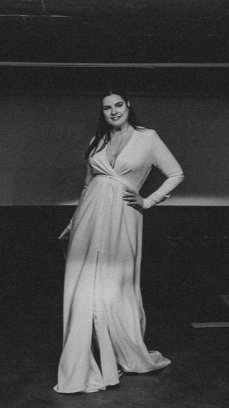 Eine Frau in einem langen, weißen Kleid posiert mit der Hand in der Hüfte in einem schwach beleuchteten Raum.