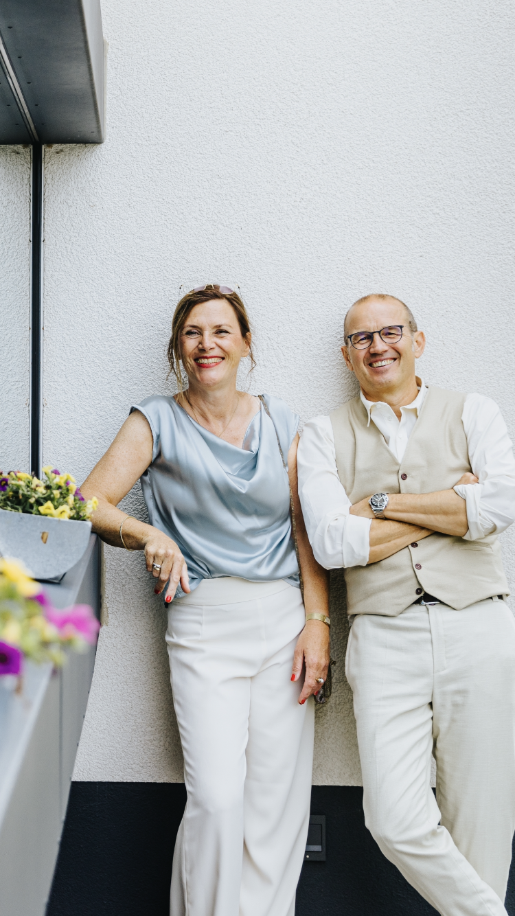 Frau in hellblauem Oberteil und Hose, Mann in Weste und Hose, lächelnd, an eine weiße Wand gelehnt.