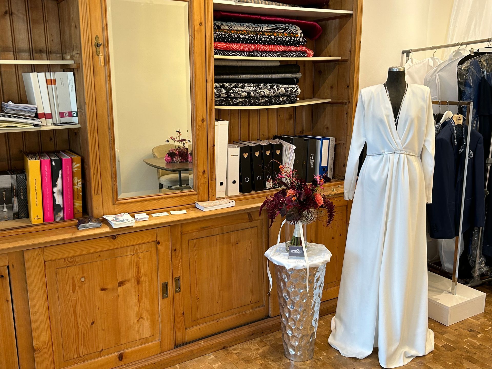 Weißes Kleid an einer Schaufensterpuppe in einem Bekleidungsgeschäft, neben einem Holzschrank mit Stoffen und Mustern.