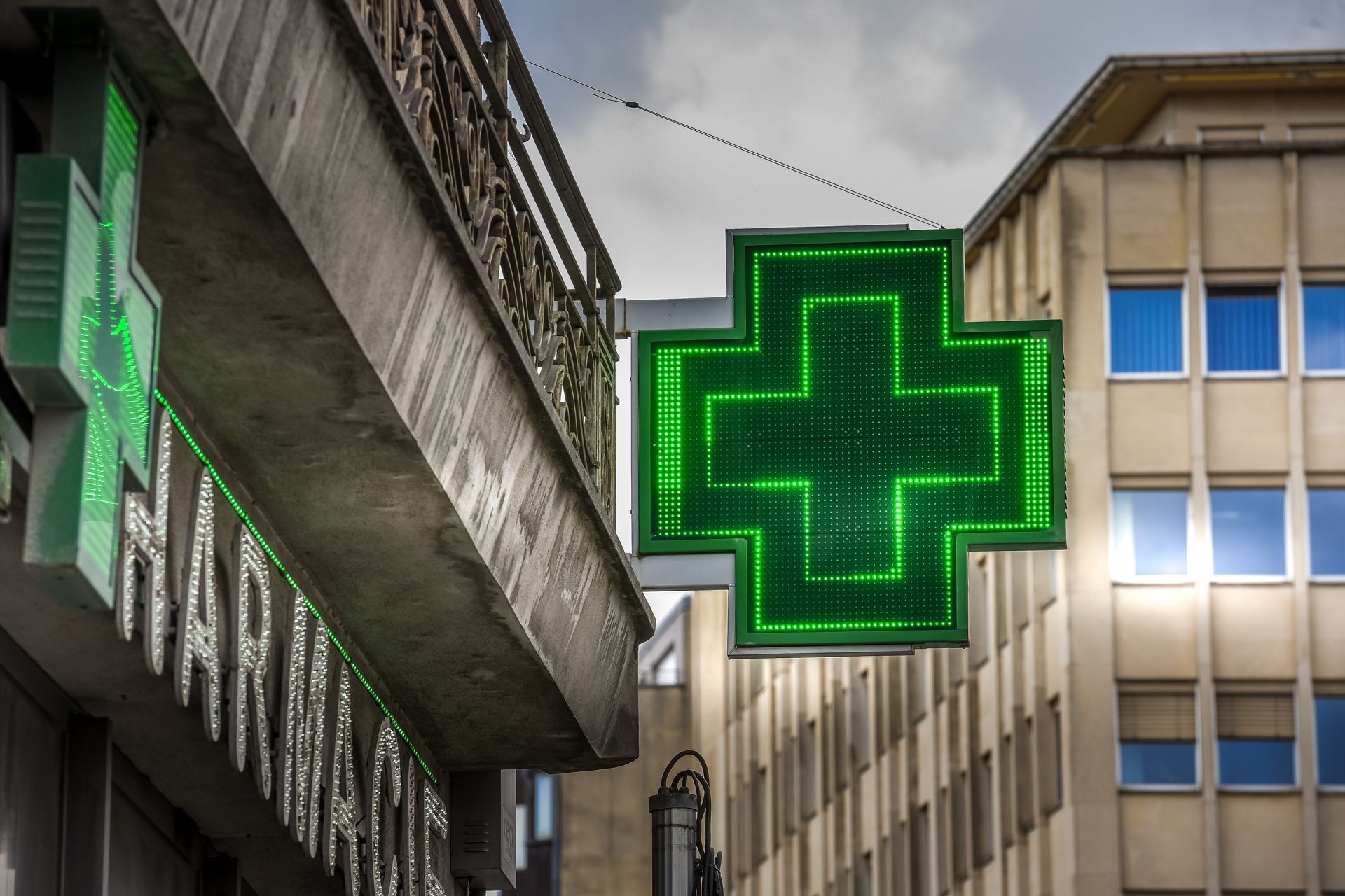 Enseigne lumineuse en forme de croix d'une pharmacie, de couleur vert fluo, suspendue entre les immeubles de la ville au niveau de la rue.