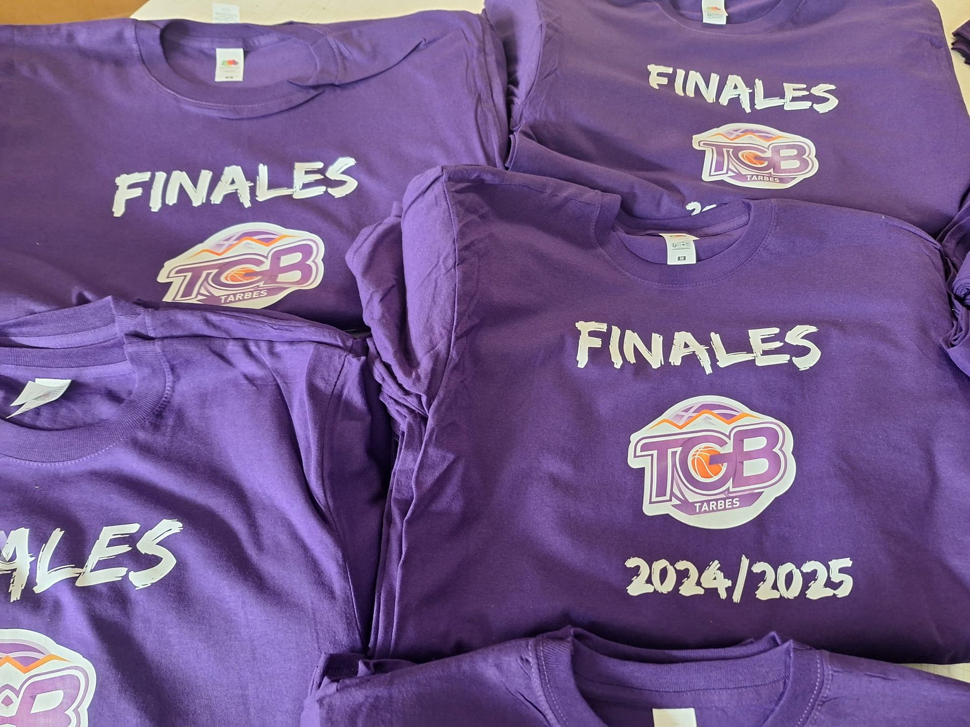 Pile de t-shirts violets imprimés FINALES et TGB en blanc et or