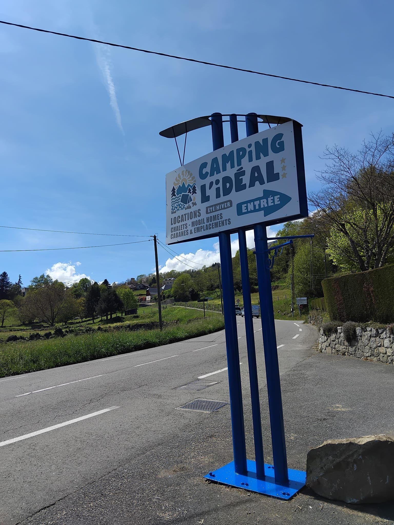 Panneau bleu indiquant un Camping au bord d'une route de campagne, au milieu de champs verdoyants sous un ciel dégagé.