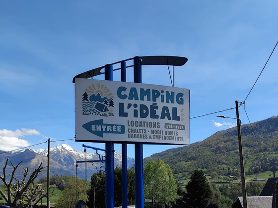 Panneau routier indiquant Camping L'Idéal sur fond de montagnes et de ciel bleu