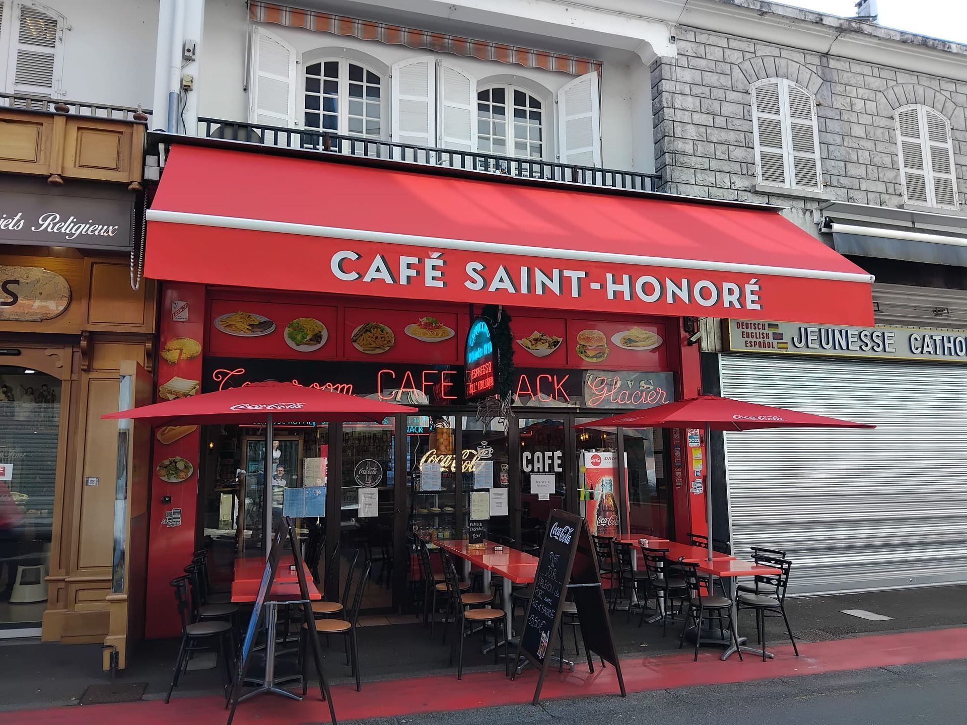Façade rouge d'un café avec auvents et tables en terrasse, portant l'inscription Café Saint-Honoré.