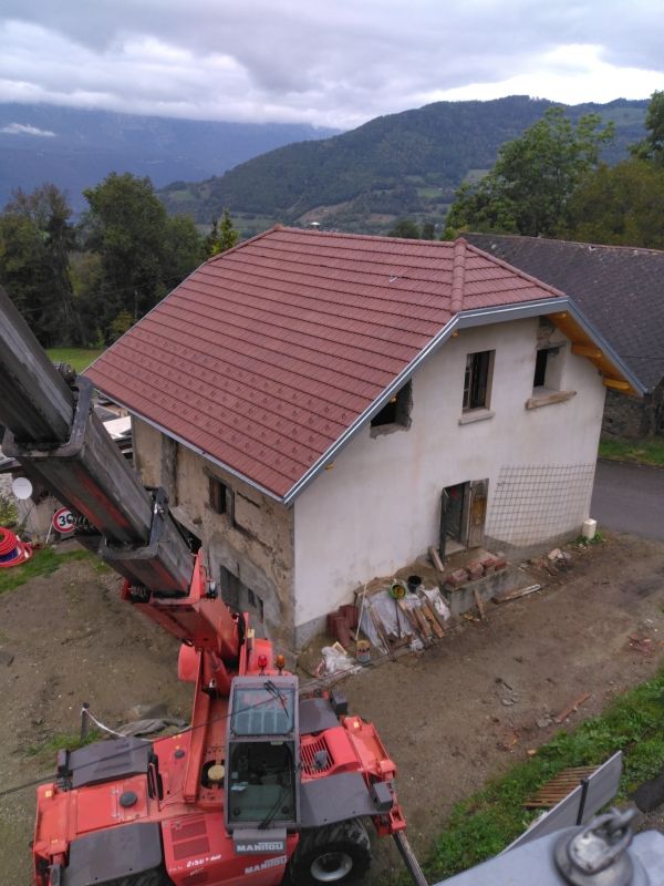 Maison au toit rouge soulevée par deux grues de chantier rouges dans un cadre rural montagneux